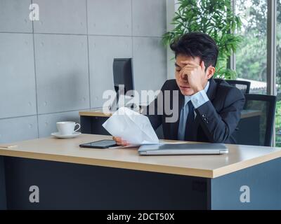 Junger asiatischer Geschäftsmann, der schlechte Nachrichten mit einem Kündigungsschreiben erhält, müde, gestresst und traurig mit abwesenden Menschen auf seinem Schreibtisch im Büro sitzt. Frustriert Stockfoto