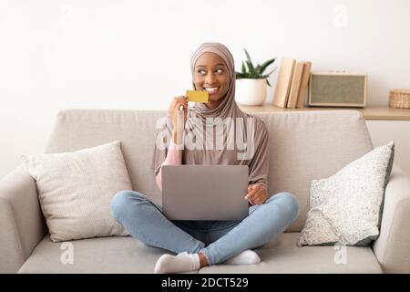 In voller Länge Porträt der schwarzen Frau in Hijab Einkaufen im Internet mit Kreditkarte und Laptop, drinnen Stockfoto