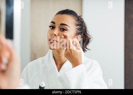 Schöne Frau im Bademantel Anwendung Anti-Aging-Creme auf ihre Wange Stockfoto