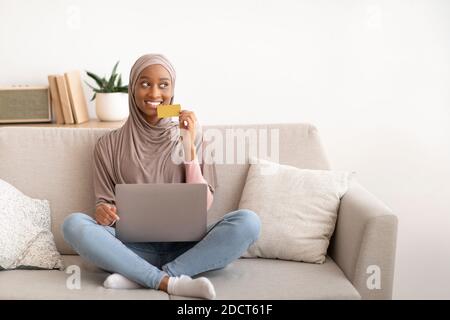 Fröhliche afroamerikanische Dame in Hijab sitzt auf dem Sofa mit Laptop und Kreditkarte, online einkaufen zu Hause, Kopierraum Stockfoto