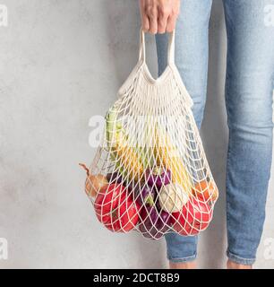 Junge Frau hält einen wiederverwendbaren Netzbeutel mit frischem Gemüse. Grauer Hintergrund, Null Abfall Konzept Stockfoto