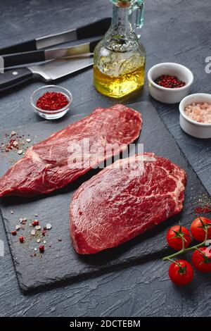 Zwei rohe Ribeye Steak, Würze, Zange und ein Messer für Fleisch auf dem Schieferbrett auf schwarzem Hintergrund. Blick von oben Stockfoto