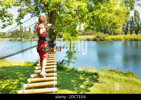 Kleiner Junge gehen auf der Seilbrücke zwischen zwei Bäume in der Nähe des Sees im Sommer Vergnügungspark Spielplatz Stockfoto