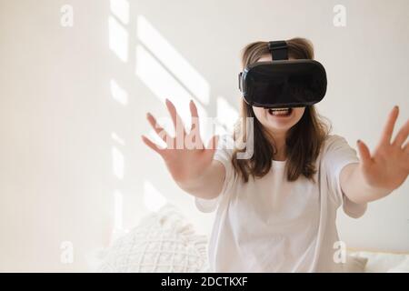 Junge Frau in Jeans und weißem T-Shirt mit Virtual Reality Brille auf dem Kopf bewegt ihre Arme, tanzt, bekommt Angst. Das Konzept der modernen Technologie, Stockfoto