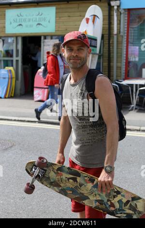 In Newquay, Cornwall, England, hält ein junger Mann sein Skateboard im geschäftigen Stadtzentrum und fängt die lebhafte Atmosphäre dieser beliebten Stadt ein. Stockfoto