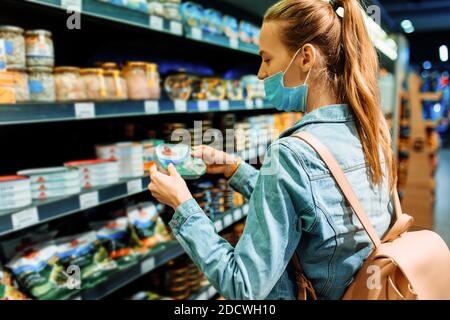 Junge Frau in einer medizinischen Schutzmaske auf ihrem Gesicht kauft Lebensmittel in einem Supermarkt. Eine junge Dame wählt Meeresfrüchte in einem Lebensmittelgeschäft. Verkauf, Kauf, Verkauf Stockfoto