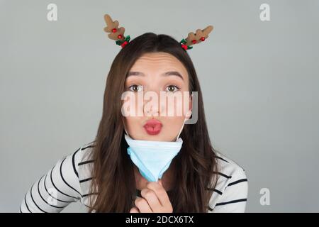 Schöne junge Brünette mit einer medizinischen Schutzmaske auf einem grauen Hintergrund. Eine Frau posiert mit einer Weihnachtsdekoration in ihren Haaren. Stockfoto