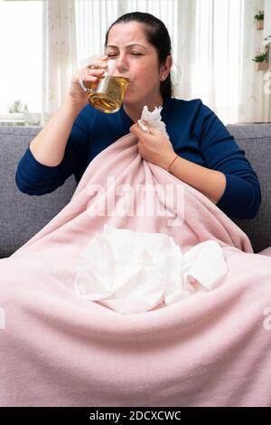 Kranke Frau zu Hause sitzt auf der Couch trinken heißen Tee Aus transparentem Glas Becher als covid Influenza Kälte Behandlung halten Servietten zum Nasenblasen Stockfoto