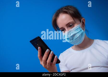 Portrait der Frau trägt medizinische Gesichtsmaske, Blick auf Smartphone in blauen Raum Stockfoto