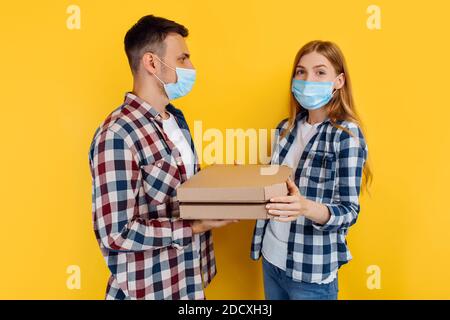 Mann und Frau in karierten Hemden und medizinischen Schutzmasken auf ihren Gesichtern, die Pizzakisten auf einem isolierten gelben Hintergrund halten. Stockfoto