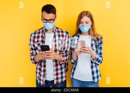 Junges Paar, ein Junge und ein Mädchen in karierten Hemden und medizinischen Schutzmasken, mit Mobiltelefonen, isoliert auf einem gelben Hintergrund Stockfoto