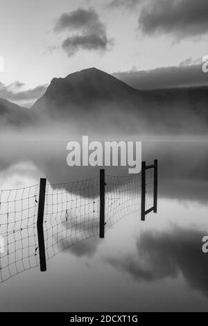 Am frühen Morgen rollt Nebel über Buttermere im englischen Lake District mit Holzzaun, der sich im Wasser spiegelt. Stockfoto