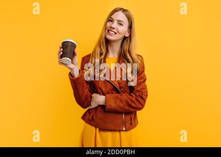 Stilvolle glücklich schöne junge Frau in einem gelben Kleid und Herbstjacke, hält eine Papiertasse Kaffee, auf einem isolierten gelben Hintergrund. Das Konzept Stockfoto