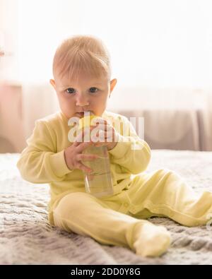 Ein brütende Baby in gelben Kleidern sitzt zu Hause mit einer Wasserflasche in den Händen. Blick auf die Kamera Stockfoto