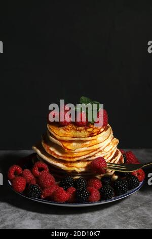 Ein Stapel hausgemachter Pfannkuchen auf einem Teller mit Himbeeren und Brombeeren auf dunklem Hintergrund. Das Konzept einer hausgemachten Mahlzeit. Stockfoto