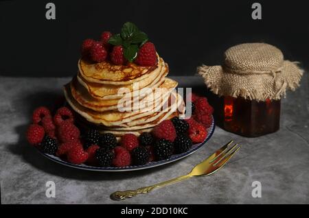 Ein Stapel hausgemachter Pfannkuchen auf einem Teller mit Himbeeren und Brombeeren und einem Glas Honig auf einem dunklen Hintergrund. Das Konzept einer hausgemachten Mahlzeit. Stockfoto