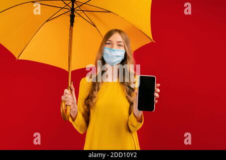 Eine schöne junge Frau in einer Schutzmaske auf ihrem Gesicht, mit einem Regenschirm in den Händen, zeigt ihr Handy Stockfoto