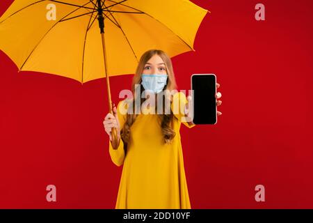 Geschocktes Mädchen in einer chirurgischen Schutzmaske auf ihrem Gesicht, in einem gelben Kleid und mit einem gelben Regenschirm in ihren Händen zeigt einen leeren Handy-Bildschirm Stockfoto