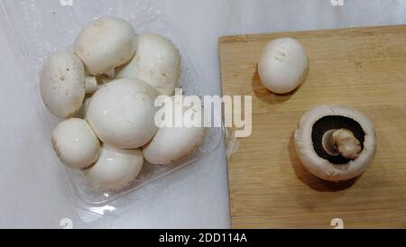 Draufsicht auf ein transparentes Plastikpaket mit weißen Champignons, mit zwei separaten Pilzen auf einem hölzernen Schneidebrett platziert. Stockfoto