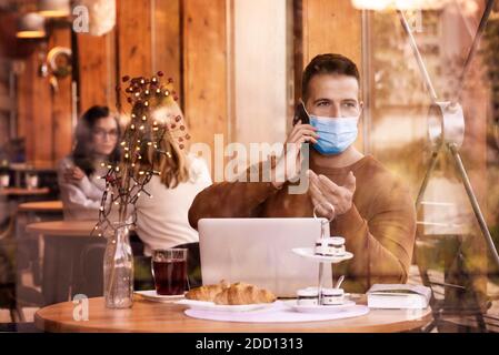 Aufnahme eines gutaussehenden Mannes mit Gesichtsmaske, während er im Café sitzt und online arbeitet. Stockfoto