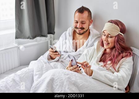 Mann und Frau ruhen sich mit dem Handy auf dem Bett aus, sie liegen morgens am Wochenende auf dem Bett, kaukasischer Mann und Frau schauen sich Videos oder etwas anderes zusammen an, schauen auf den Bildschirm Stockfoto