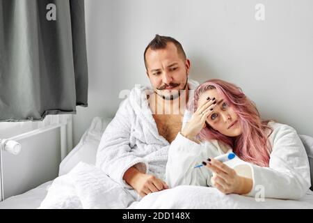 Kranke Frau mit Ehemann im Bett, Weibchen hat hohe Temperatur, leidet an Fieber. Im Schlafzimmer Stockfoto
