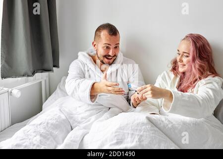 Glückliches kaukasisches Paar, das mit positivem Schwangerschaftstest am Morgen auf dem Bett aufgeregt wird, glückliche Reaktion, ist Mann überrascht Stockfoto