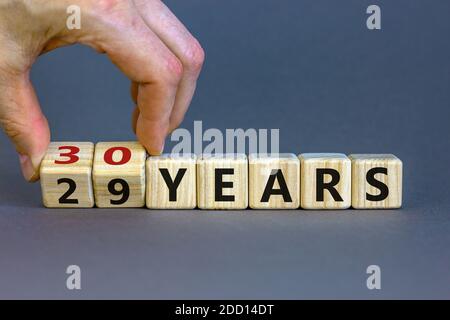 Männliche Hand dreht Würfel und ändert den Ausdruck "29 Jahre" zu "30 Jahre" oder umgekehrt. Schöner grauer Hintergrund. Geschäfts- und Feierkonzept, Stockfoto