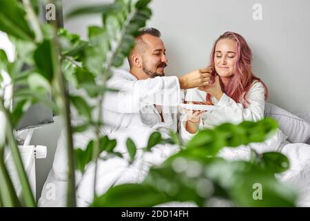 Paar behandeln einander mit eclars auf dem Bett, haben Frühstück zusammen, schöne Frau und Mann in Bademänteln genießen, entspannen Sie am Morgen. Blick durch grüne Pflanzen Stockfoto