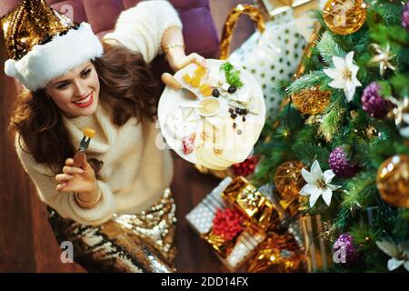 Frohe Weihnachten. Obere Ansicht der lächelnden mittleren Alters Hausfrau in Gold Pailletten Rock und Pullover mit großen Käseplatte in der Nähe Weihnachtsbaum und Geschenkboxen Stockfoto