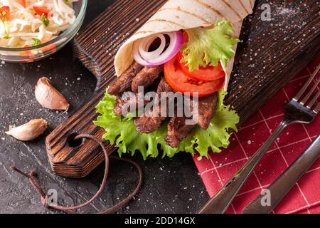 Handmade Wrap Shawarma liegt auf dem Holz Schneidebrett. Shawarma wird mit Tortilla, Rindfleisch, Tomaten, Zwiebeln und Salat auf einem schwarzen Tisch hergestellt. Stockfoto