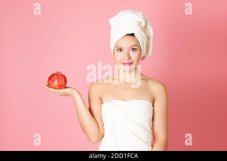 Eine schöne junge Frau mit einem Handtuch auf dem Kopf und Körper hält einen Granatapfel auf Armlänge, isoliert auf einem rosa Hintergrund Stockfoto