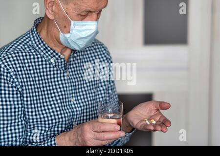 Älterer kranker Mann in einer medizinischen Maske. Er hält Pillen und ein Glas Wasser in den Händen. Stockfoto