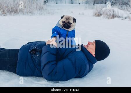 Mops Hund spielt auf Schnee mit seinem Besitzer auf dem Boden liegen. Welpe trägt Wintermantel im Park. Kleidung für Tiere Stockfoto