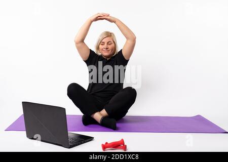 Ältere Frau meditiert auf der Matte vor dem Laptop und hält die Hände über dem Kopf. Stockfoto