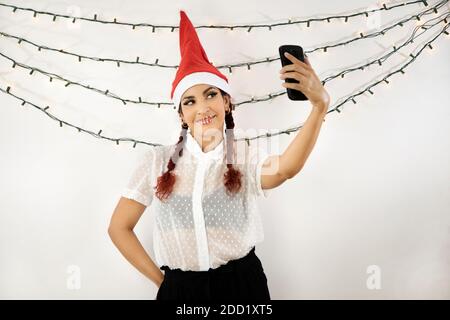 Mädchen mit einem roten Weihnachtshut und roten Haaren, mit einem Handy, das einen Videoanruf macht, lächelnd ein Selfie-Foto macht. Schönes Mädchen Stockfoto