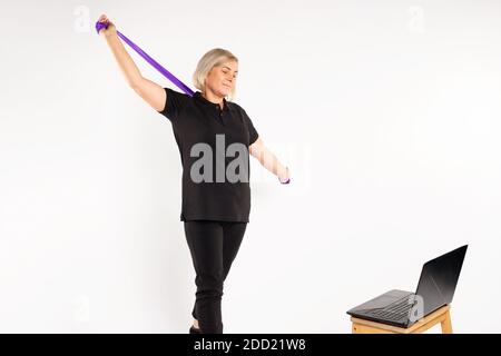Sportliche ältere Frau, weißer Hintergrund dehnt Fitnessband vor dem Laptop. Stockfoto