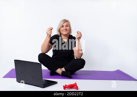 Eine ältere Frau sitzt mit gekreuzten Beinen auf dem Boden vor einem Laptop und meditiert. Stockfoto