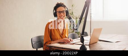Frau, die Podcast mit Mikrofon und Laptop aufzeichnet. Weibliche Podcasterin, die auf die Kamera schaut und lächelt, während sie von zu Hause aus arbeitet. Stockfoto