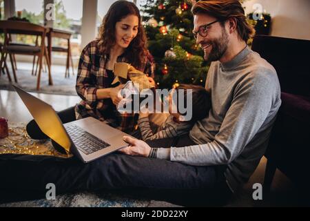 Mann mit Videoanruf auf seinem Laptop mit seinem Sohn und Frau Eröffnung Weihnachtsgeschenke. Familie feiert Weihnachten zu Hause während Pandemie, und verbunden Stockfoto