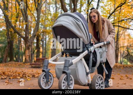 Fröhliche Mutter spielt mit neugeborenen Baby im Kinderwagen. Frau, die mit Kinderwagen im Park spazieren geht Stockfoto