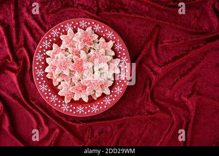 Schneeflocken-förmige Zuckerkekse mit roten Streuseln, auf einer roten Platte mit weißen Schneeflocken, rote Tischdecke Stockfoto