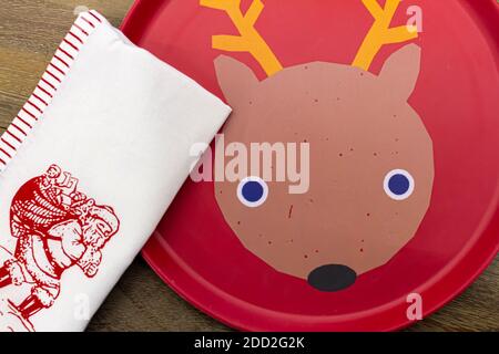 Ein roter Einwegteller mit einer lustigen Rein-Hirsch-Figur im Inneren wird auf einem Holztisch platziert. Eine weiße Stoffserviette mit Weihnachtsmann-Bild ist plac Stockfoto
