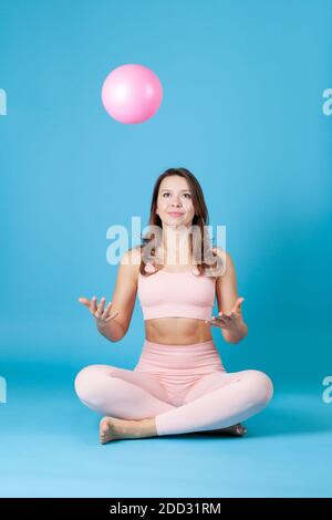 Eine junge Frau in einem rosa Sporttop und Leggings sitzt mit gekreuzten Beinen und wirft einen Ball auf, isoliert auf einem blauen Hintergrund Stockfoto