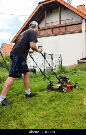 Teenager mäht einen Rasen in einem Hinterhof in Potzbach, Deutschland, während sein Hund hinter einem Zaun wacht. Stockfoto