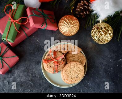 Leckeres Shortbread mit Schokoladenstücken auf einem kleinen Teller, Spielzeug auf dem Weihnachtsbaum, Fichtenspieß, Geschenke, Weihnachtsmütze, Blick von oben. Flach Stockfoto