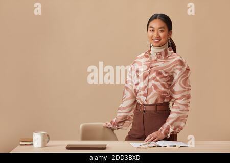 Porträt einer jungen asiatischen Geschäftsfrau, die lächelt und die Kamera in der Nähe steht Ihr Arbeitsplatz vor dem beigen Hintergrund Stockfoto