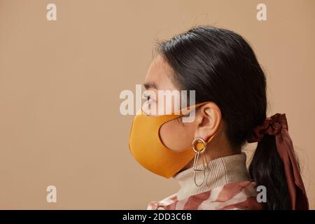 Seitenansicht der asiatischen jungen Frau trägt Schutzmaske stehen Vor dem beigen Hintergrund Stockfoto