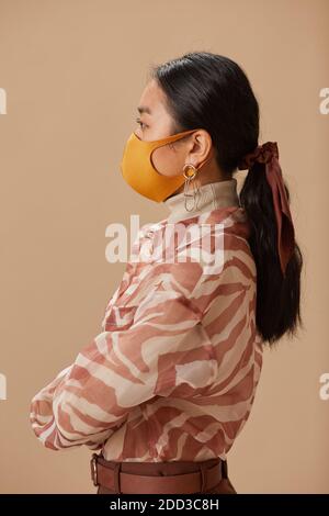 Seitenansicht der asiatischen jungen Frau in eleganten Kleidung tragen Schutzmaske isoliert auf beigem Hintergrund Stockfoto
