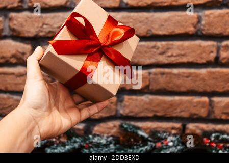 Geschenkkarton mit einer roten Schleife in der Hand auf dem Hintergrund einer Ziegelwand und einem Weihnachtsbaum Zweig. Stockfoto